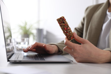 Woman holding tasty granola bar working with laptop at light table in office, closeupの写真素材