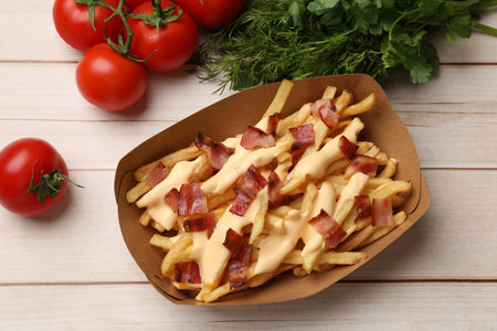 Tasty potato fries, cheese sauce, bacon and products in paper container on light wooden table, top...