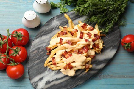 Delicious French fries with bacon, cheese sauce, tomatoes and herbs on light blue wooden table, flat layの写真素材