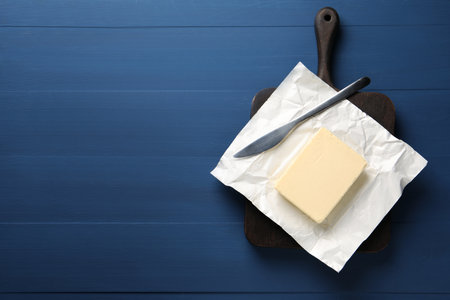 Block of tasty butter in open foil packaging and knife on blue wooden table, top view. Space for textの写真素材