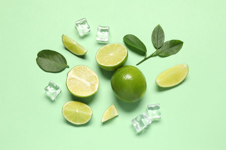 Fresh ripe limes with leaves and ice cubes on light green background, flat layの写真素材