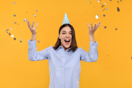 Happy young woman in party hat near flying confetti on yellow backgroundの写真素材