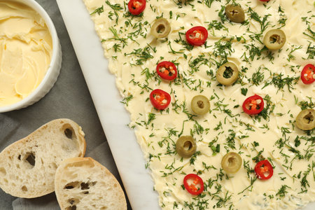 Fresh butter board with cut olives, pepper and bread on gray tablecloth, flat lay