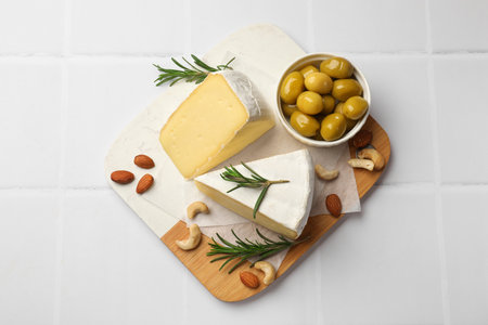 Pieces of tasty camembert cheese, rosemary, nuts and olives on white tiled table, top viewの写真素材