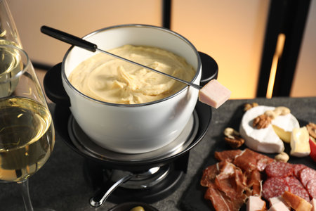 Fork with piece of ham, fondue pot with melted cheese, wine and snacks on gray table, closeupの写真素材