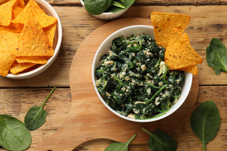 Tasty spinach dip with eggs in bowl and nachos chips on wooden table, flat lay