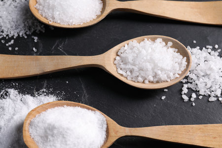 Natural salt and wooden spoons on black table, flat lay