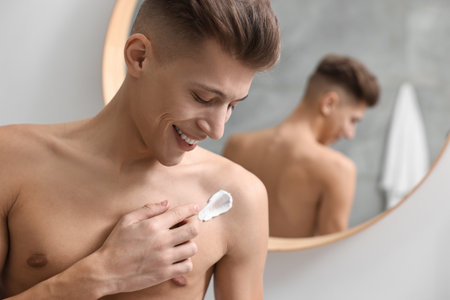 Handsome man applying moisturizing cream onto his shoulder indoors. Space for textの写真素材
