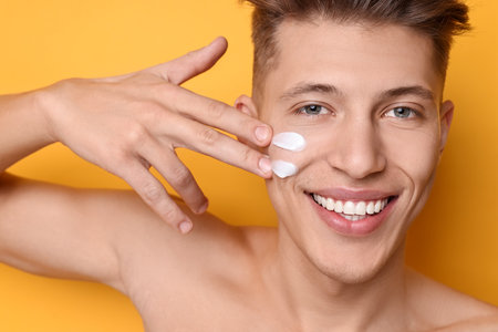 Handsome man applying moisturizing cream onto his face on orange background, closeupの写真素材