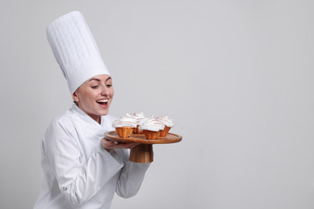 Happy professional confectioner in uniform holding delicious cupcakes on light grey background. Space for textの写真素材