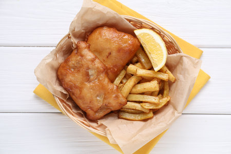 Tasty fish, chips and lemon in wicker bowl on white wooden table, top view