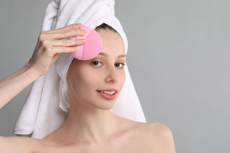 Washing face. Young woman with cleansing brush on grey background, space for textの写真素材