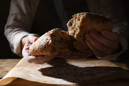 Man breaking loaf of fresh bread at wooden table, closeup