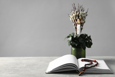 Burning candle, plant with willow branches, Bible and rosary beads on gray table, space for text
