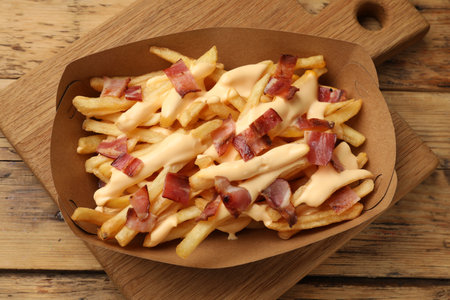 Tasty potato fries, cheese sauce and bacon in paper container on wooden table, top viewの写真素材