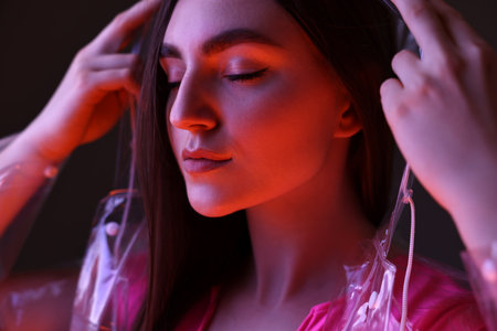 Fashionable portrait of beautiful woman wearing transparent coat on dark background in neon lights,...