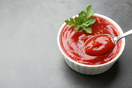 Taking delicious tomato ketchup with spoon from bowl at gray textured table, closeup. Space for textの写真素材