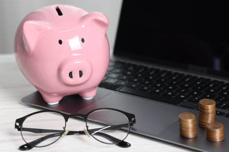Piggy bank, glasses, coins and laptop on white tableの写真素材