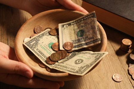 Donate and give concept. Woman holding bowl with money at wooden table, closeup
