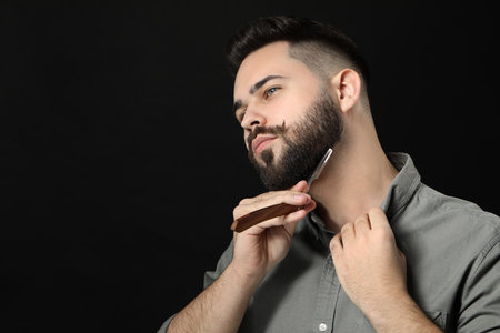 Handsome young man shaving beard with blade on black background. Space for textの写真素材