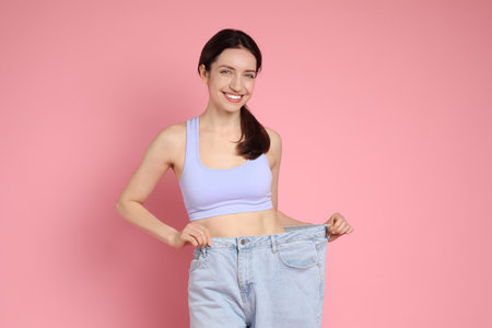 Happy young woman in big jeans showing her slim body on pink backgroundの写真素材