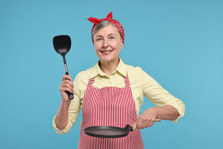 Happy housewife with turner and frying pan on light blue backgroundの写真素材