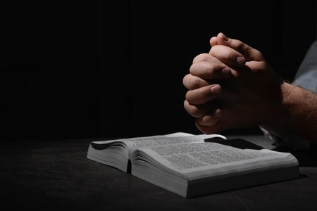 Religion. Christian man praying over Bible at table against black background, closeup. Space for textの写真素材