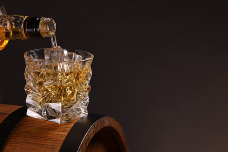 Pouring whiskey from bottle into glass on wooden barrel against dark background, closeup. Space for textの写真素材