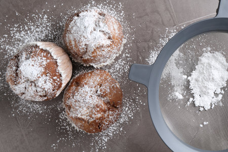 Sieve with sugar powder and muffins on gray textured table, flat lay