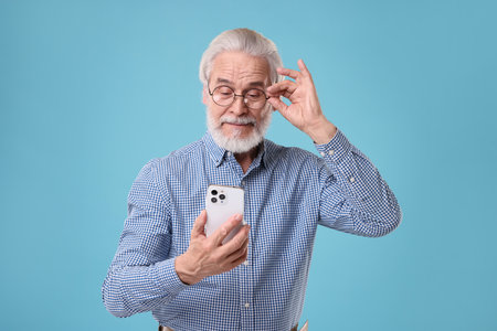 Portrait of stylish grandpa with glasses using smartphone on light blue backgroundの写真素材