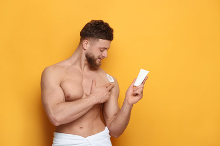 Handsome man applying moisturizing cream onto his shoulder on orange background, space for textの写真素材