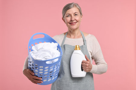 Happy housewife with detergent and basket full of laundry on pink backgroundの写真素材