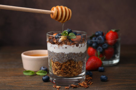 Pouring honey onto tasty granola with berries, yogurt and chia seeds at wooden tableの写真素材