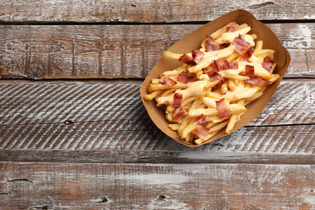 Tasty potato fries, cheese sauce and bacon in paper container on wooden table, top view. Space for...