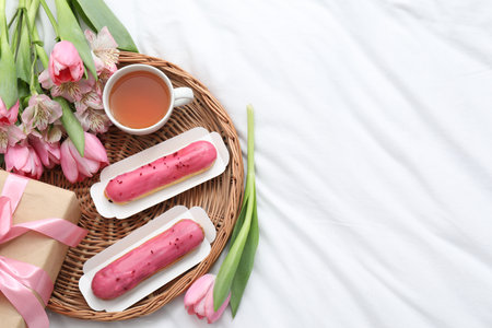 Tasty breakfast. Delicious eclairs, tea, flowers and gift box on bed, top view. Space for textの写真素材