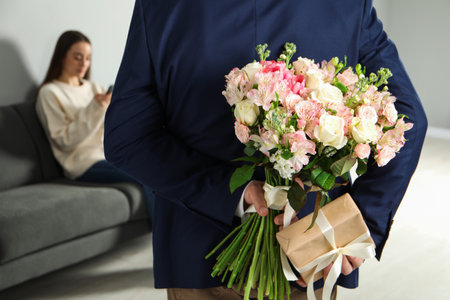 Man hiding bouquet of flowers and present for his beloved woman indoors, closeupの写真素材