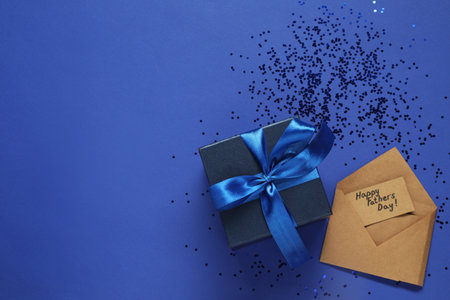Card with phrase Happy Father's Day, gift box and confetti on blue background, flat lay. Space for...