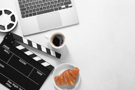 Movie clapper, film reel, laptop, coffee and croissant on light textured table, flat lay. Space for...