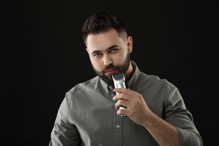 Handsome young man trimming beard on black background