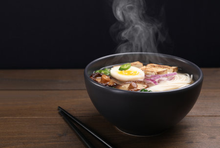 Delicious hot vegetarian ramen in bowl on wooden table against black background, closeup. Space for...