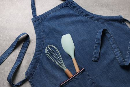 Stylish blue apron, spatula and whiskey on gray table, top view