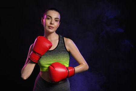 Portrait of beautiful woman wearing boxing gloves in color lights on black background. Space for...