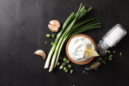 Delicious yogurt in bowl, green onion, garlic and salt on black background, flat lay. Space for textの写真素材