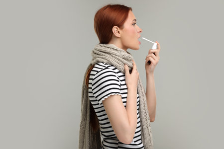 Young woman with scarf using throat spray on grey backgroundの写真素材