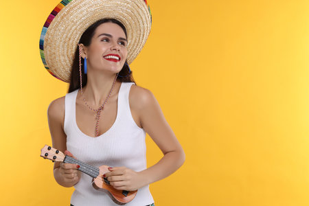 Young woman in Mexican sombrero hat playing ukulele on yellow background. Space for textの写真素材