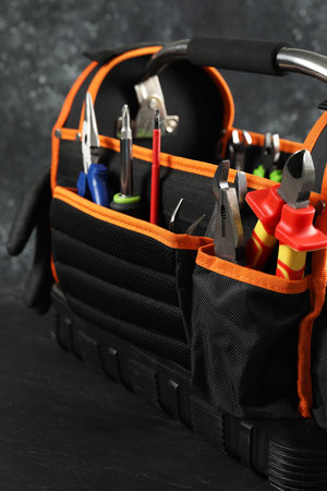 Bag with different pliers, wrench and other tools for repair on black table, closeup
