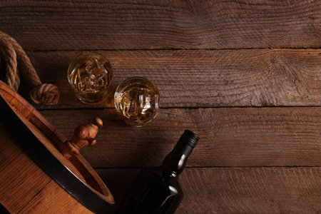 Whiskey with ice cubes in glasses, bottle and barrel on wooden table, flat lay. Space for textの写真素材