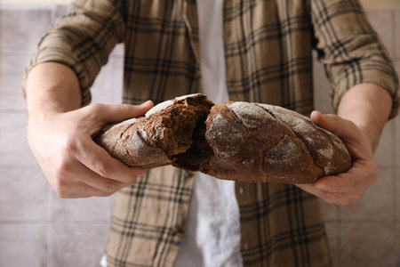 Man breaking loaf of fresh bread near gray wall, closeup