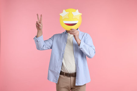 Man holding emoticon with stars instead of eyes and showing peace sign on pink backgroundの写真素材