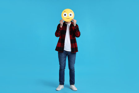 Man covering face with surprised emoticon on light blue backgroundの写真素材